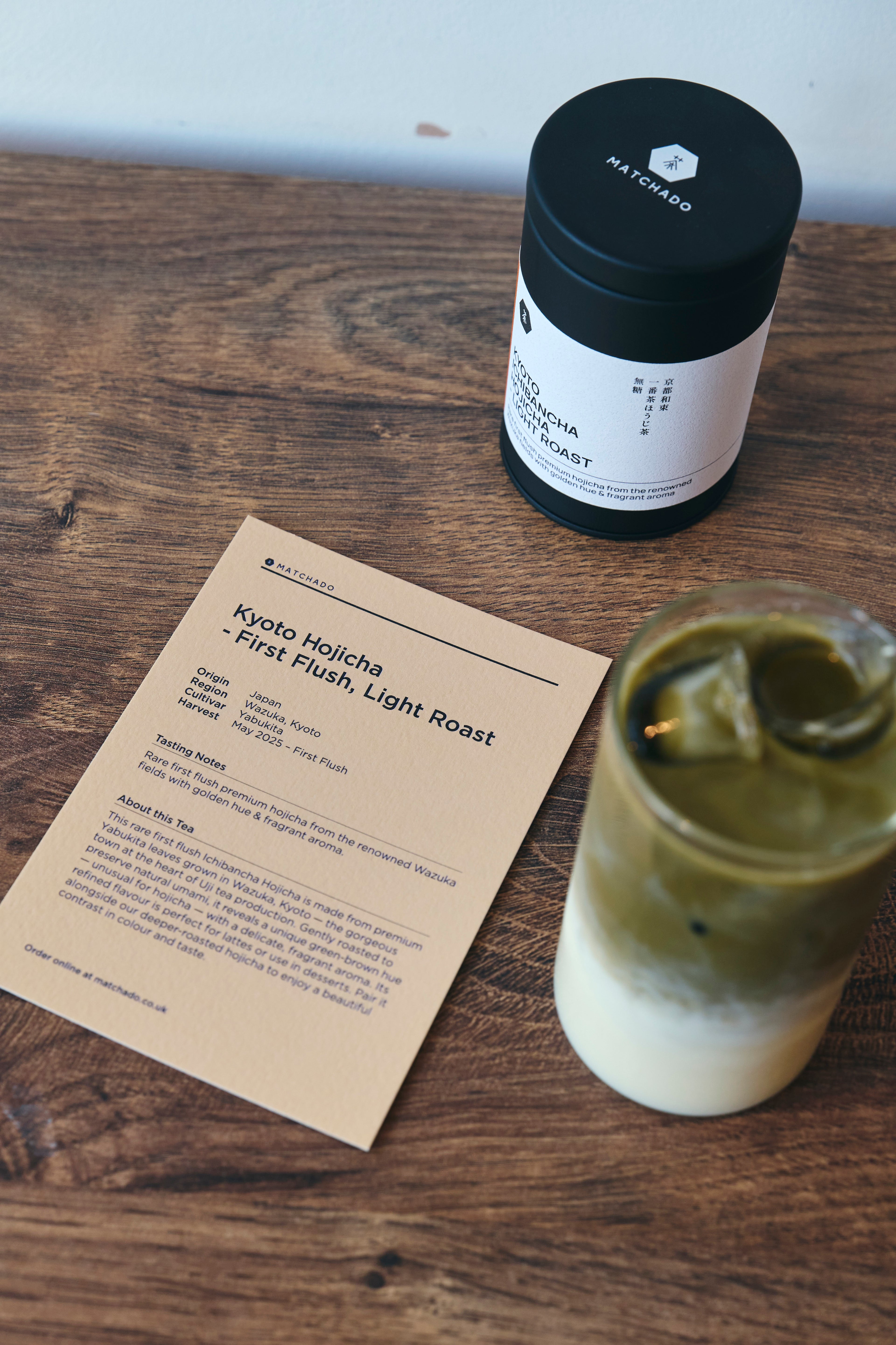 MATCHADO Flush Kyoto Hojicha Powder Jar of Kyoto Hojicha tea, a paper bag with text, and a glass with a green liquid on a wooden surface.