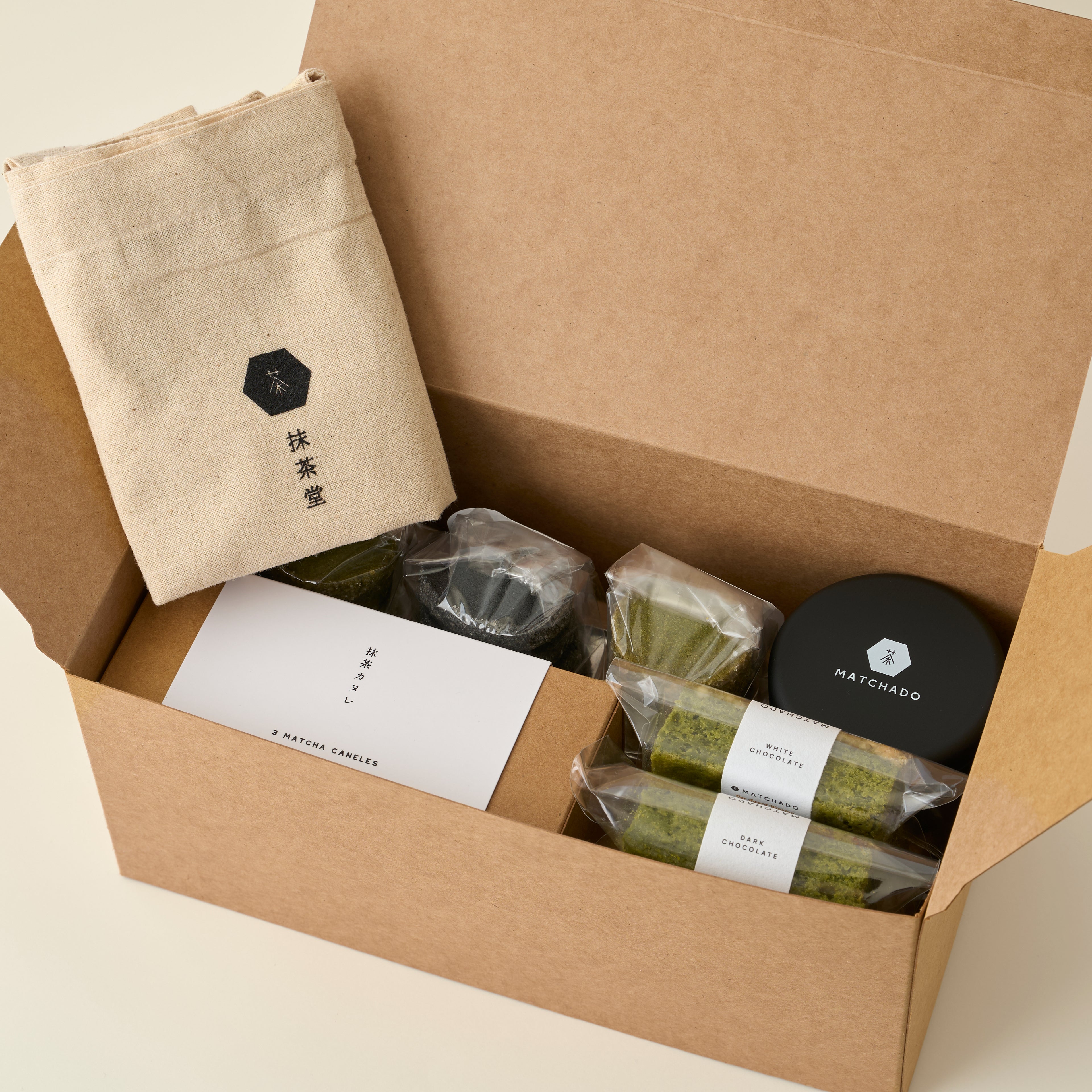 MATCHADO Afternoon tea set. Open cardboard box containing matcha green tea products with a beige cloth and black container.