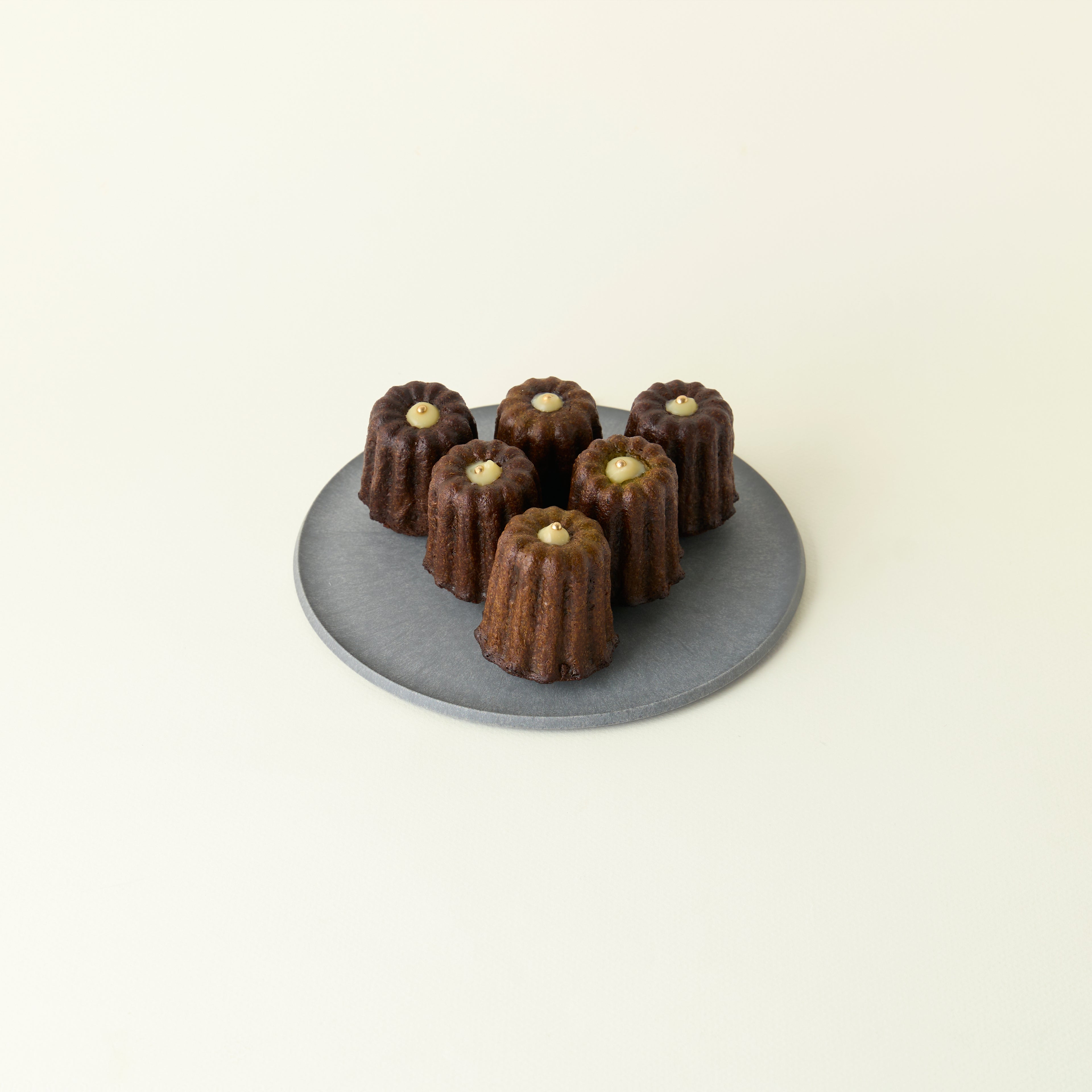 Six chocolate canelés on a gray plate against a beige background