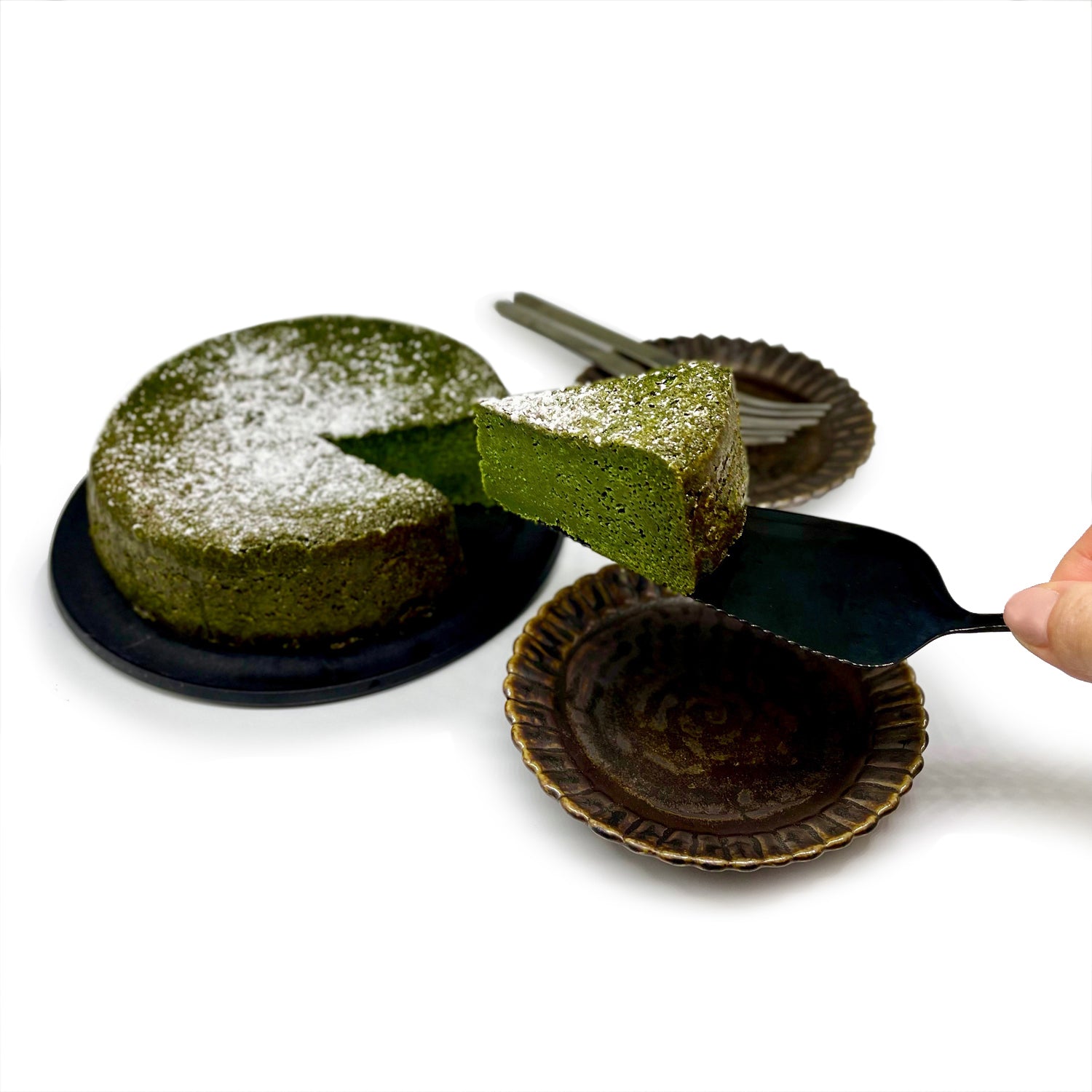 A slice of MATCHADO Matcha Gâteau Chocolat being served with a sleek black cake server. The photo displays the rich green texture of the cake alongside the whole gâteau.