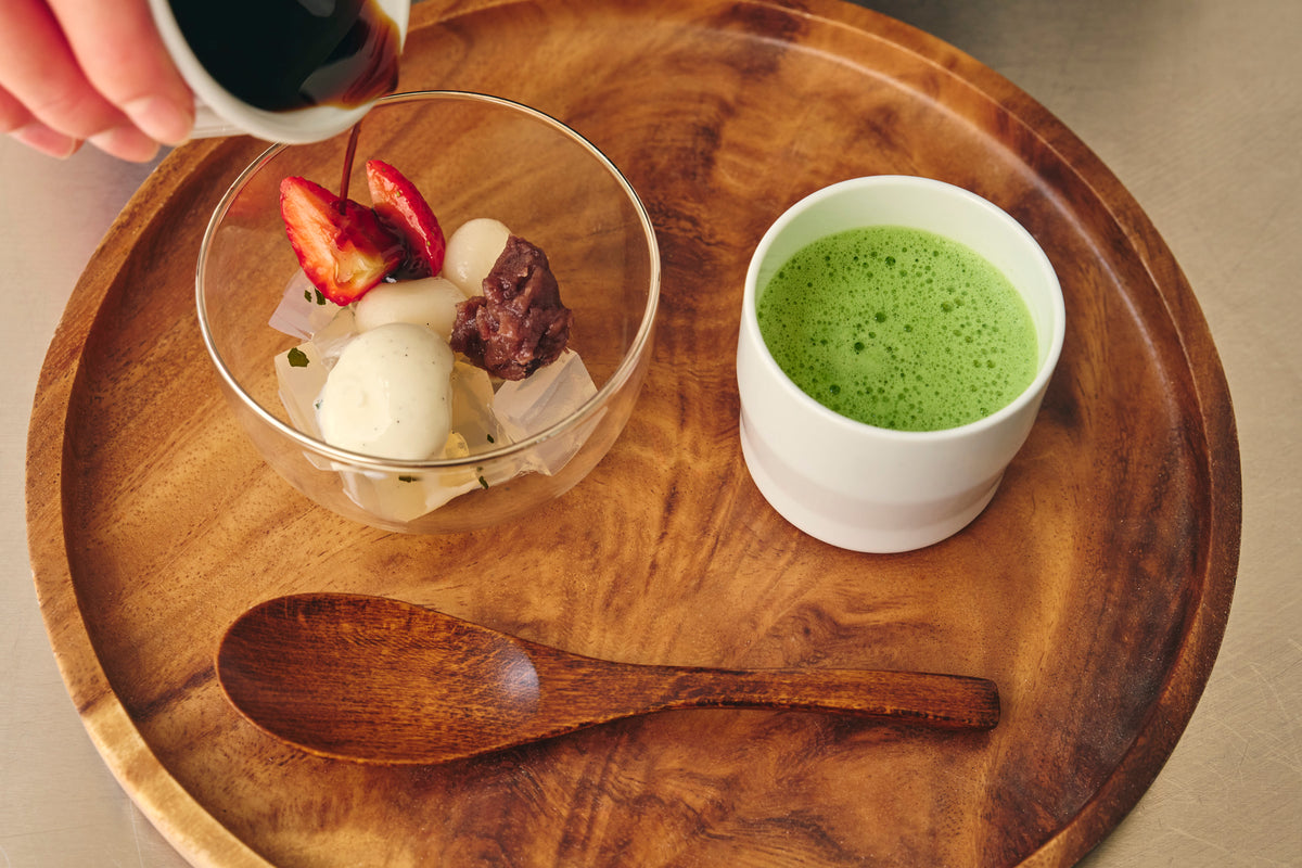 A styled food scene on a round wooden tray with a glass bowl containing dessert pieces and a white cup of bright green matcha drink. A hand is pouring brown sugar syrup from a small white pitcher into the bowl, with a wooden spoon resting below.