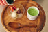 A styled food scene on a round wooden tray with a glass bowl containing dessert pieces and a white cup of bright green matcha drink. A hand is pouring brown sugar syrup from a small white pitcher into the bowl, with a wooden spoon resting below.