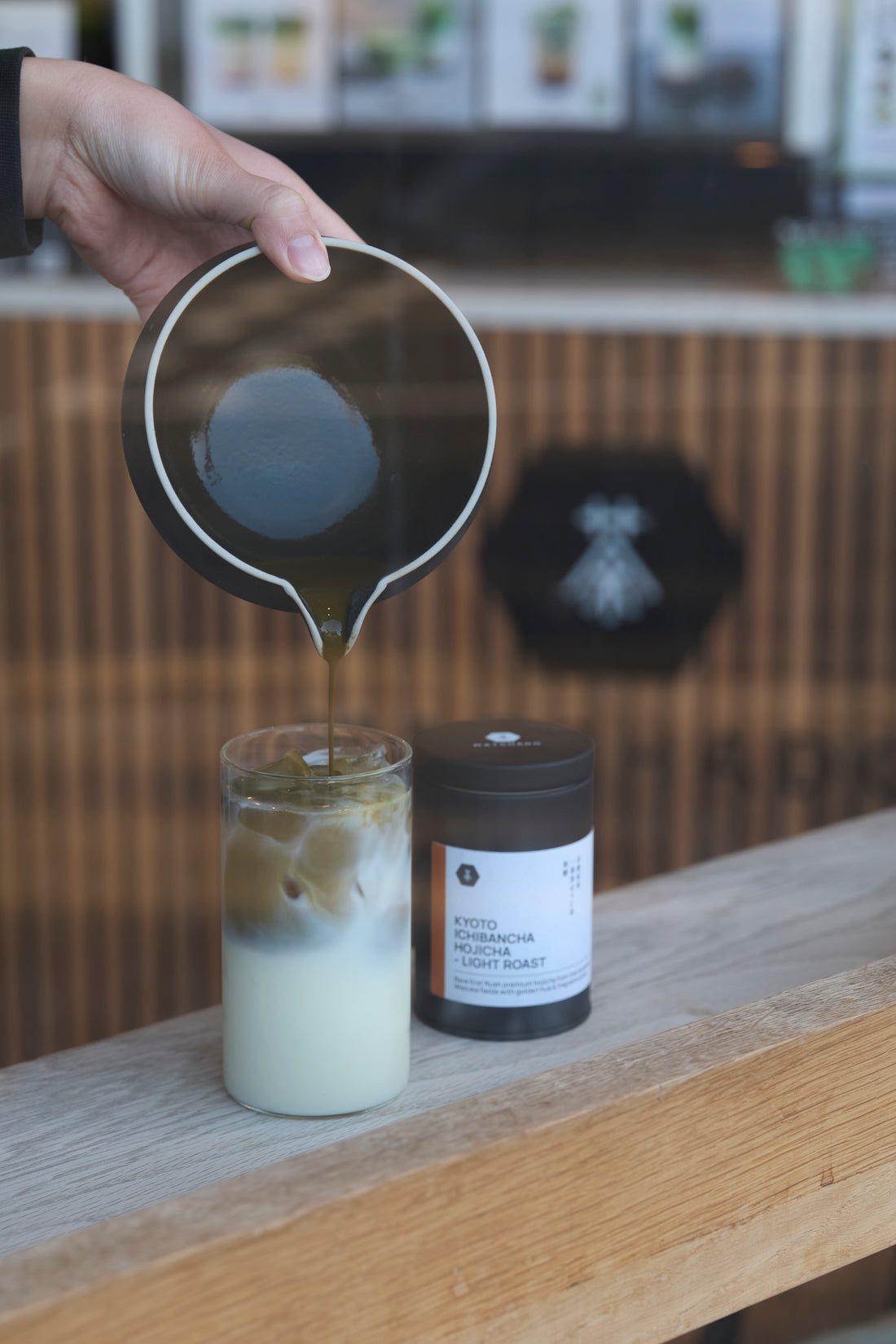 MATCHADO Flush Kyoto Hojicha Powder. Hand pouring a liquid into a glass with a blurred coffee shop background