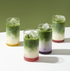 MATCHADO four glasses of layered green and white drinks with ice cubes on a white surface.