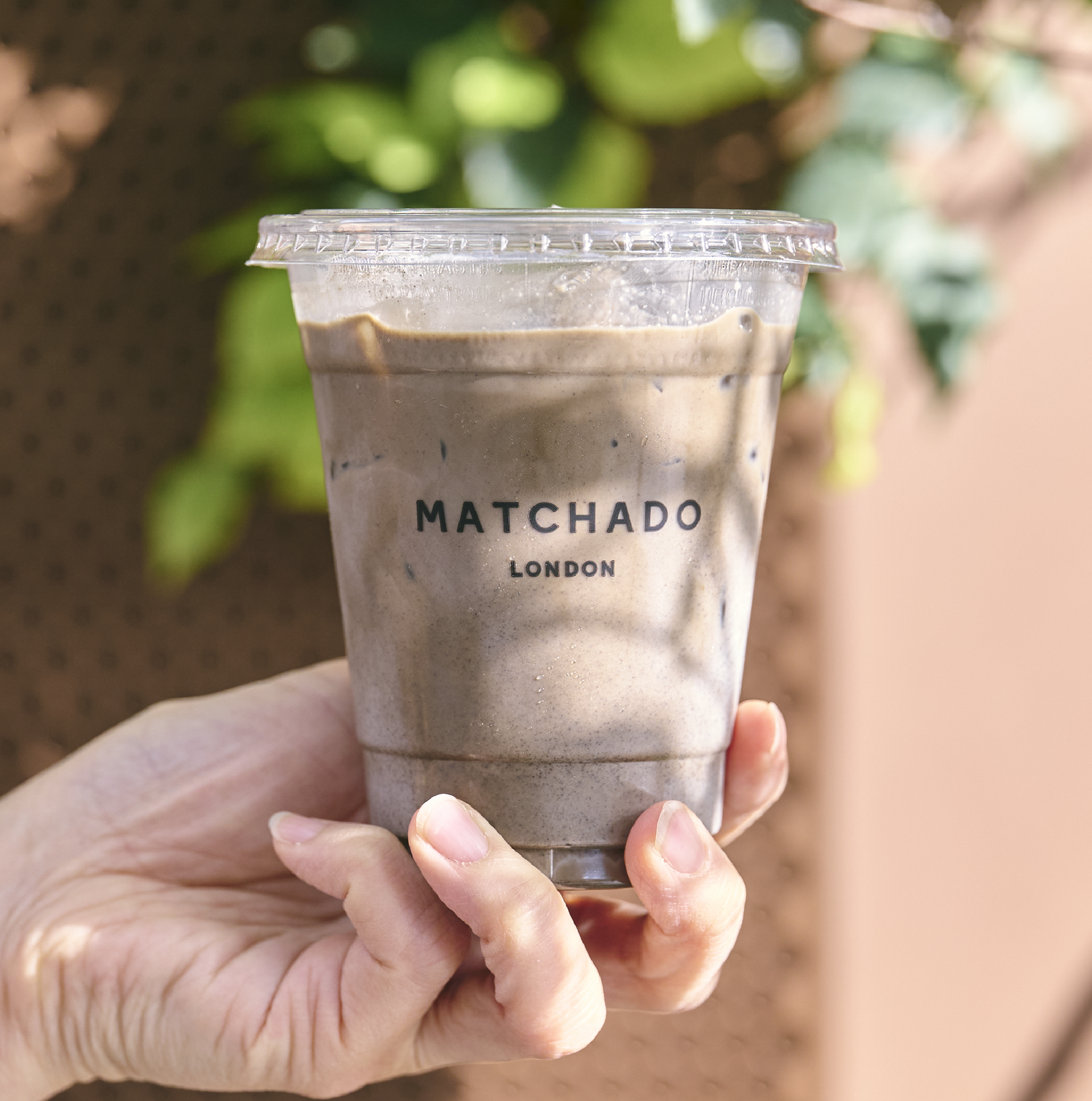 Hand holding a cup of Matchado London iced Hojicha latte with a blurred green background