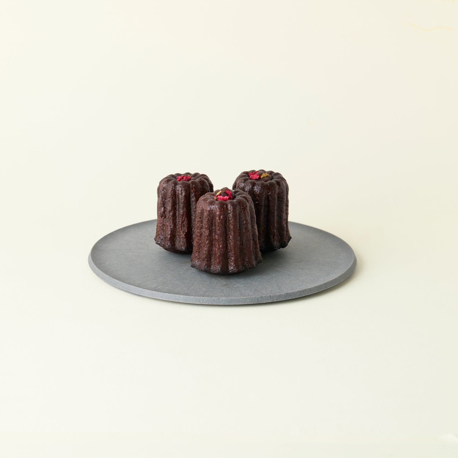 Three Chocolate Canelés on a grey plate, topped with dried raspberries and gold leaf. The dark, caramelized crust reflects a MATCHADO twist on the classic French pastry.