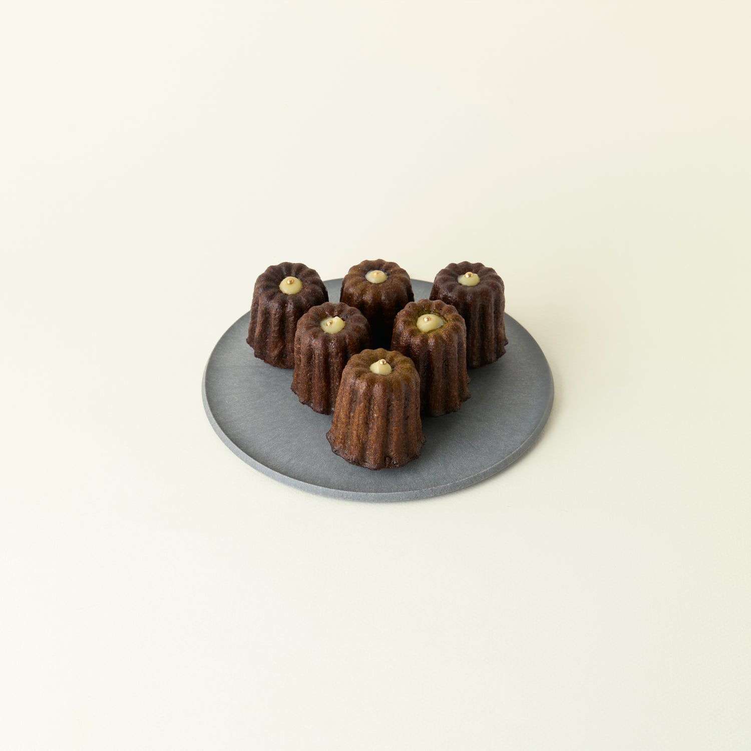 Six chocolate canelés on a gray plate against a beige background