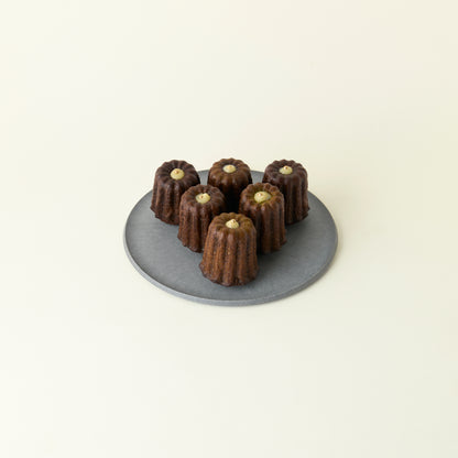 Six chocolate canelés on a gray plate against a beige background