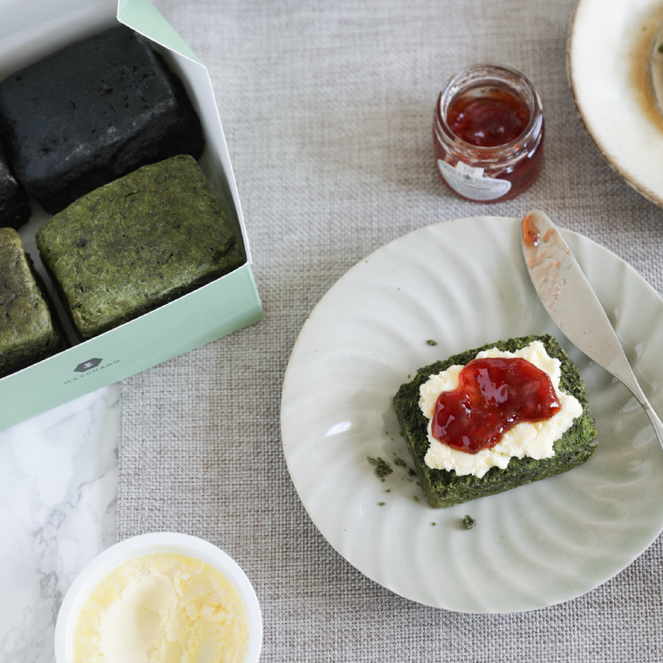 MATCHADO Matcha Scones with cream and jam on a plate, next to a box of green tea cakes and a jar of jam.